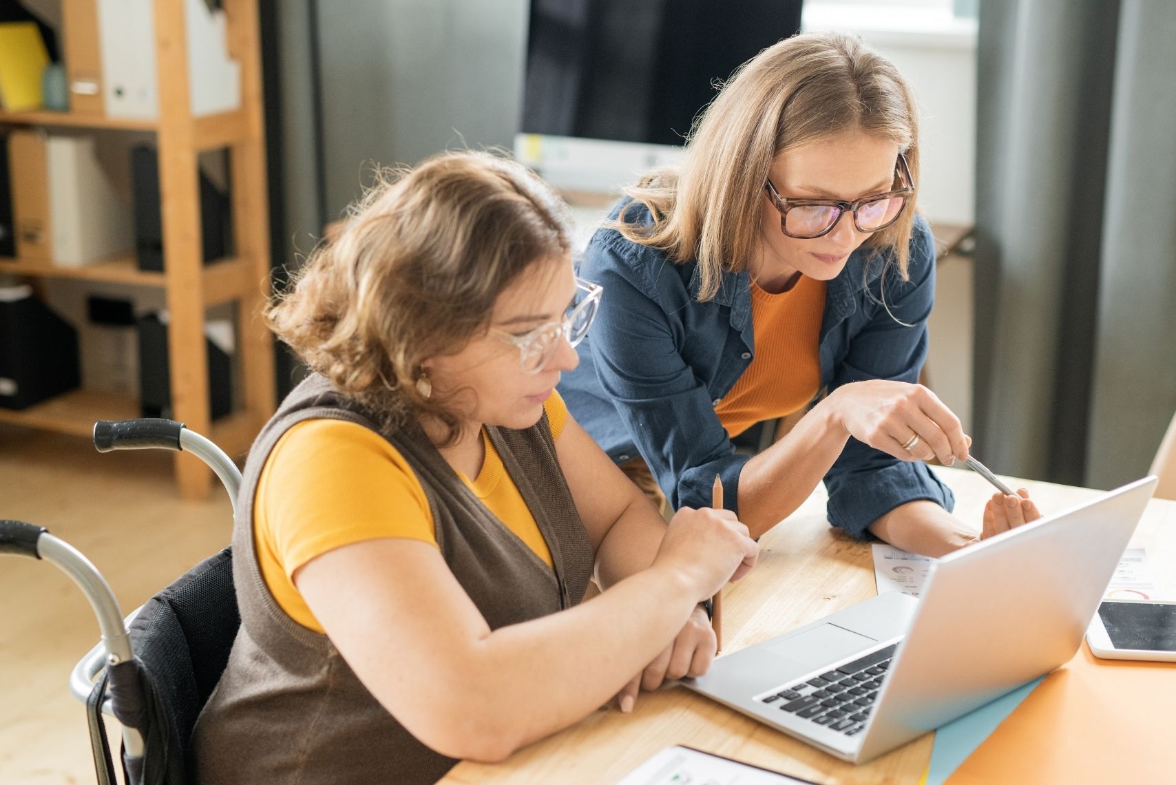 Two women in wheelchairs collaborate on a laptop, focused on their work in a bright, modern office setting.